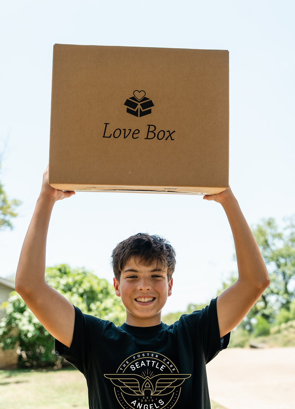 Alt text: Smiling boy wearing a Seattle Angels T-shirt holds a large cardboard “Love Box” overhead outdoors on a sunny day