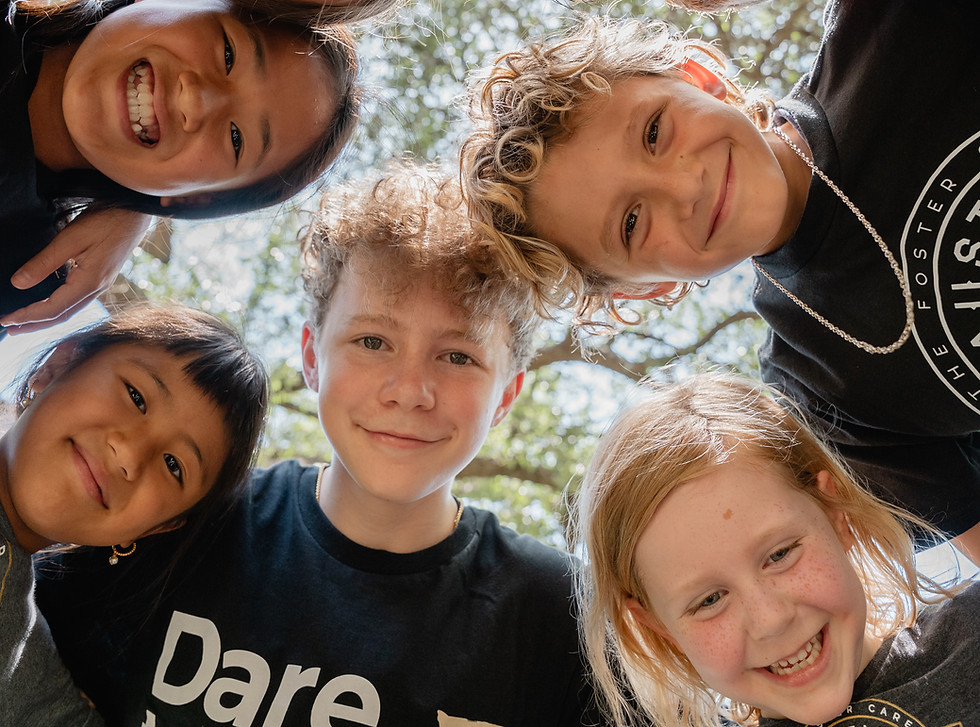 Five smiling children lean in close outdoors, looking down at the camera in a circle beneath sunlit tree branches